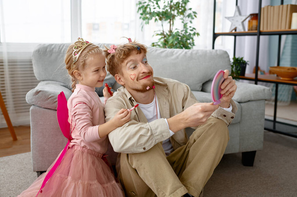 Маленькая счастливая дочь и отец с забавным макияжем, любящая семья проводит время дома - Фото, изображение