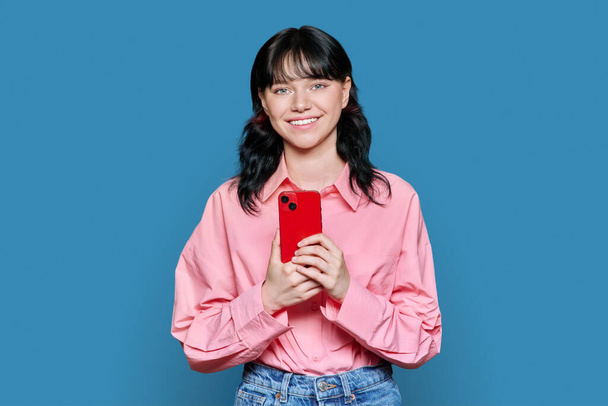 Young teenage girl with smartphone looking at camera on blue background. Smiling female student using mobile phone. Text message social network online communication, mobile applications leisure study - Foto, immagini