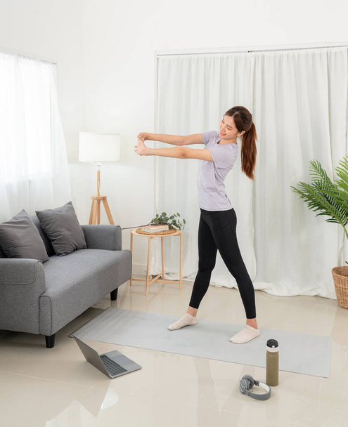 Молода жінка бере урок йоги онлайн на ноутбуці і тренує вправи з йоги, стоячи, щоб розтягнути руки на килимку у своїй вітальні вдома
. - Фото, зображення