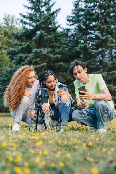 Teen-Blogger sitzen im Sommerpark und wählen trendige Tanzvideos für den Blog der sozialen Netzwerke aus. Video-Livestream. Vielschichtiges Team. Kreatives Team - Foto, Bild