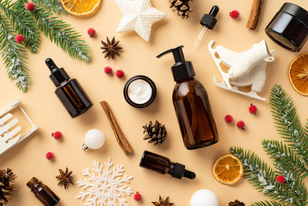 Winter skincare concept. Top view photo of amber bottles cream jar christmas ornaments white baubles mistletoe berries fir branches pine cone cinnamon dried orange slices on isolated beige background - Photo, Image