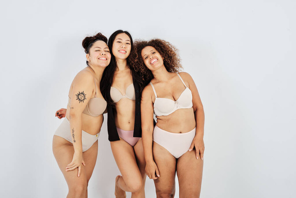 Happy multiracial female friends in lingerie smiling while standing together against gray background - Foto, Imagem