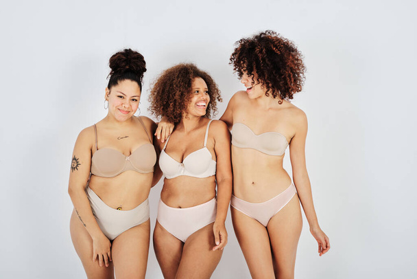 Positive multiethnic female models in lingerie standing together against gray background in studio - Foto, imagen