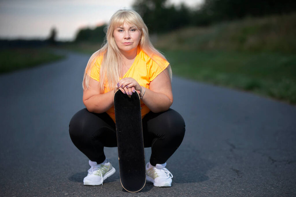 Chubby middle aged European woman, overweight, in a tracksuit with a skateboard, tired after playing sports, resting. - Photo, Image