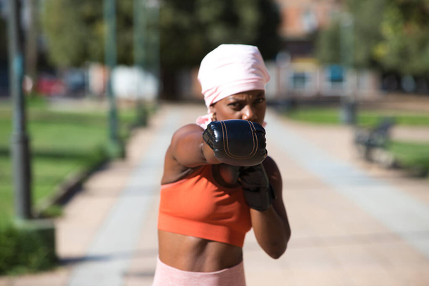 Mulher afro-americana a praticar desporto no parque. A mulher tem luvas de boxe e um cachecol cor-de-rosa na cabeça. Conceito de câncer de mama e saúde. - Foto, Imagem