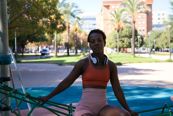 Fiatal és gyönyörű afro-amerikai nő, faragott testtel a parkban ülve edzés előtt. Narancssárga felsőben és harisnyanadrágban van, és fehér fejhallgatóval hallgat zenét. Sport és egészség. - Fotó, kép