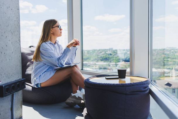 A young woman in glasses sits on a soft ottoman near the window and looks at the city from a skyscraper. Remote work - Photo, Image