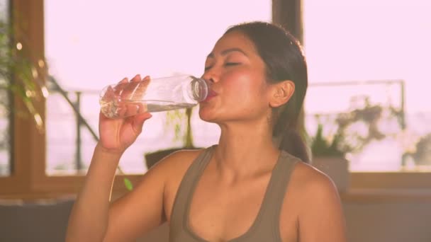 CHIUDI: Bella donna asiatica che beve acqua dalla bottiglia dopo un allenamento intensivo. Giovane donna che si prende una pausa durante l'attività sportiva domestica per idratarsi. Bere acqua rinfrescante per superare la sete. - Filmati, video