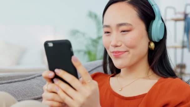 Asian woman, music headphones and smartphone social media app, podcast and audio ebook listening on living room sofa. Young girl face, mobile radio streaming and watch online technology at home. - Footage, Video