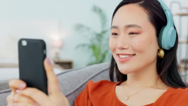 Telefoon, muziek en sociale media met een Aziatische vrouw die een bericht typt tijdens het beluisteren van audio op een koptelefoon in haar huis. Podcast, streaming en radio met een jonge vrouw die geniet van een online afspeellijst. - Video