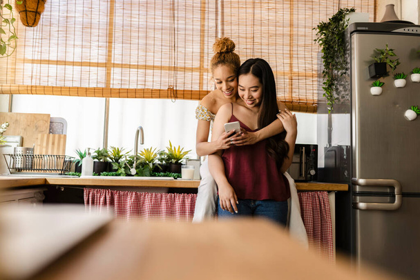 Wielorasowa para lesbijek przytulająca się w kuchni oglądając social media na smartfonie. Koncepcja LGBTQ. - Zdjęcie, obraz