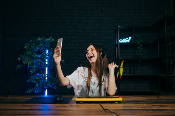 Pozitif oyuncu bir kadın bilgisayarda online bir oyun izlerken selfie çeker, gülümser ve akıllı telefon kamerasına bakar.. - Fotoğraf, Görsel