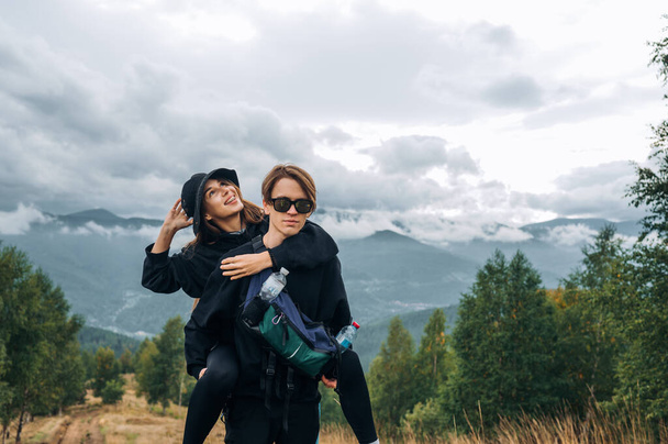Mujer feliz y hombres excursionistas en ropa casual elegante que se divierten en las montañas, el chico levantó a su novia en su espalda. - Foto, imagen