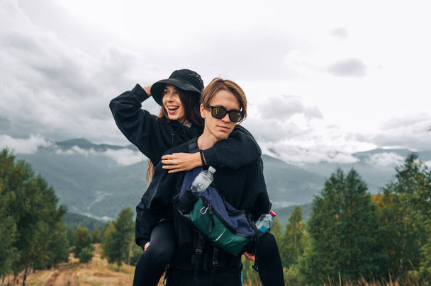 明るい若い観光客のスタイリッシュなカップルが山の上で楽しんでいる、女性は笑っている、男は美しい山の景色を背景に立って、彼の背中に彼女を運んでいる. - 写真・画像