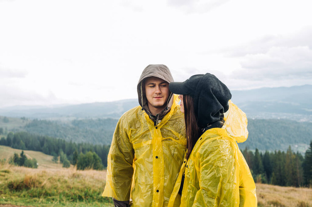 Una hermosa pareja joven en una caminata se encuentra en las montañas sobre el fondo de hermosas vistas, vestidas con impermeables y mirándose con una cara seria.. - Foto, Imagen