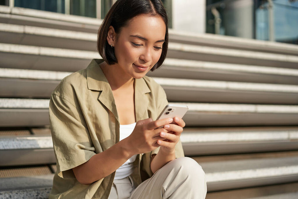 Personas y tecnología. Sonriendo hermosa mujer asiática sentada en las escaleras de la ciudad, sosteniendo el teléfono móvil. Chica con teléfono inteligente descansa fuera. - Foto, imagen