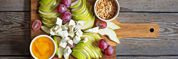 Sliced Blue cheese, dor blue or roquefort and fruits (grapes, pears), nuts and honey on a board. Exquisite appetizer for wine. Fruity cheese plate, banner.  - Фото, изображение