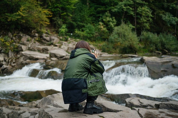 La parte posterior de una turista se sienta en una piedra en la orilla de un río de montaña y toma una foto en una cámara de teléfono inteligente - Foto, imagen