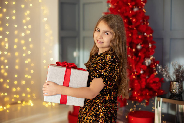 Щаслива маленька усміхнена дівчинка з різдвяною подарунковою коробкою від ялинки у затишній вітальні будинку. Красива дівчина в золотій сукні тримає різдвяний подарунок в яскравому новорічному інтер'єрі
 - Фото, зображення