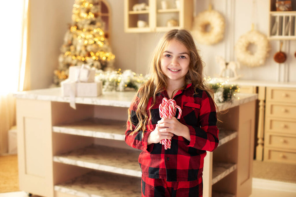 Happy young girl with a lollipops near  Christmas tree in kitchen. Little girl with sweet candies on Xmas eve. Family, tradition, celebration concept. Child girl holding festive Candy canes in hand - Photo, Image
