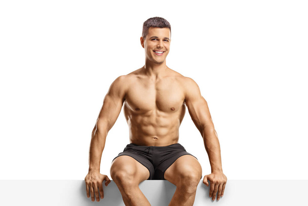 Bodybuilder sitting on a blank panel isolated on white background - Fotó, kép