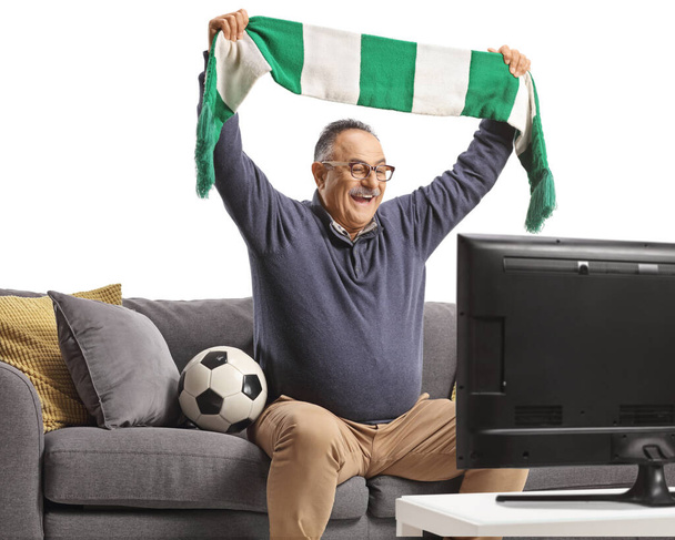 Mature man sitting on a sofa watching a football match and cheering with a scarf isolated on white background - Fotografie, Obrázek