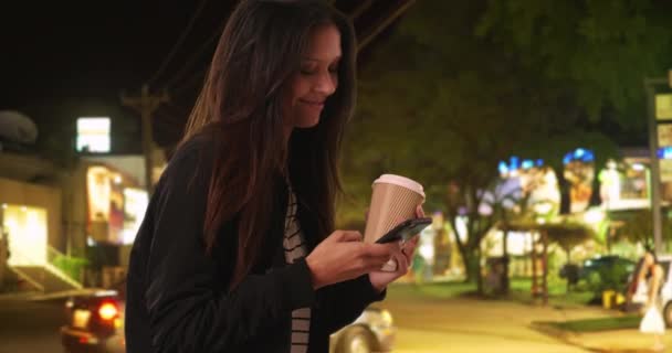 Hipster girl in bomber jacket laughs at text message from friend back home while vacationing in Costa Rica. Millennial woman with coffee on Costa Rican street at night texting with mobile phone. 4k - Footage, Video