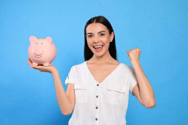 Emotional young woman with piggy bank on light blue background - Foto, Bild