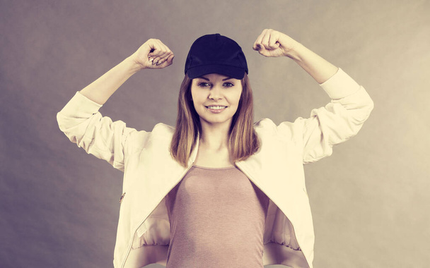 Молодая спортсменка в кепке и спортивной одежде демонстрирует мышцы рук, наслаждаясь результатами тренировок. Студия на сером фоне
 - Фото, изображение