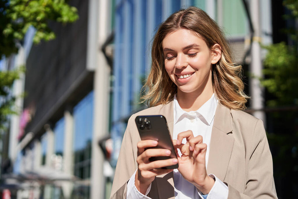 Porträt einer lächelnden schönen Geschäftsfrau, die ihr Handy auf der Straße benutzt. Corporate Frau tippt auf ihr Smartphone, Nachrichten, steht auf der Straße. - Foto, Bild