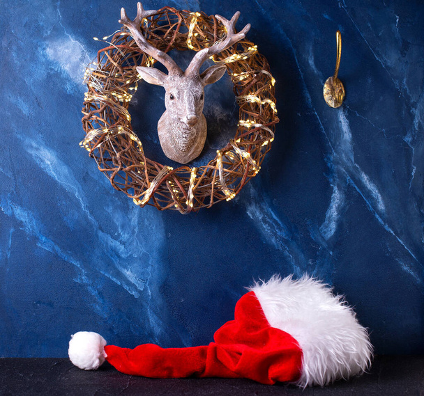 Symbols of Christmas - Santa Claus hat, wreath with fairy lights and head of deer on dark blue murmur background. Place for text. Posrtcard. - Фото, зображення