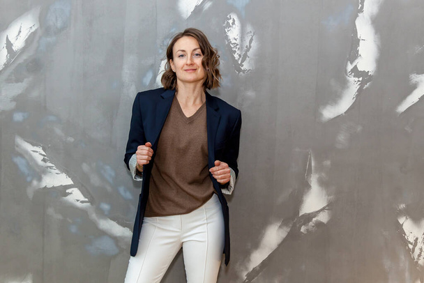 Fashionable and stylish girl posing in white trousers, a brown sweater and a black jacket against a gray wall - Fotó, kép