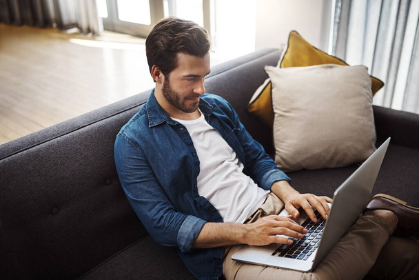 Nie denerwuj się dzisiaj i załatw sprawy z domu. przystojny młody mężczyzna korzystający z jego laptopa podczas relaksu na kanapie w domu - Zdjęcie, obraz