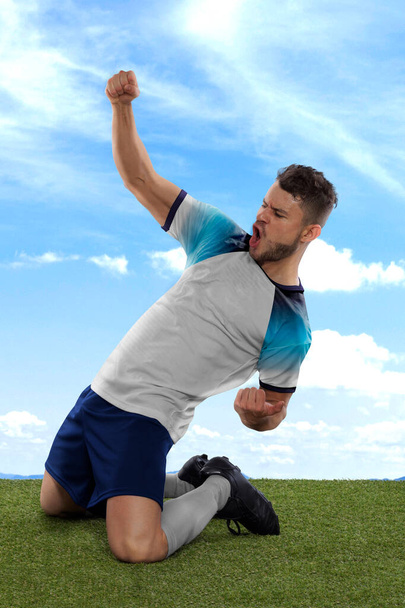 Professional soccer player with a white England national team jersey shouting with excitement for scoring a goal with an expression of challenge and happiness on field grass and cloud background - Фото, изображение