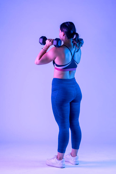 νεαρή γυναίκα σε αθλητικά, από πίσω κάνει πιεστήριο ώμων με δύο αλτήρες - Φωτογραφία, εικόνα