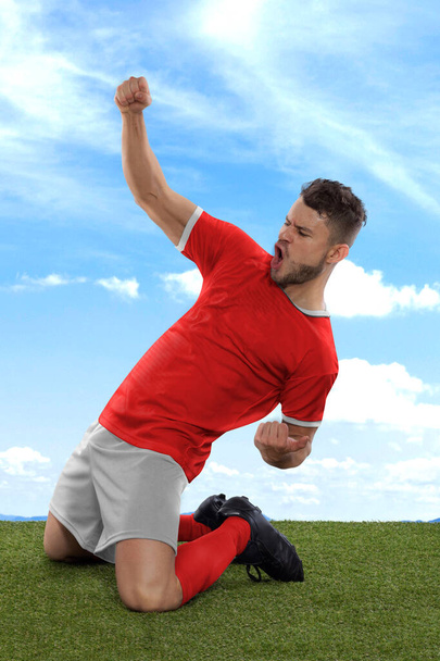 Professional soccer player with a red Tunisia national team jersey shouting with excitement for scoring a goal with an expression of challenge and happiness on field grass and cloud background - Foto, imagen