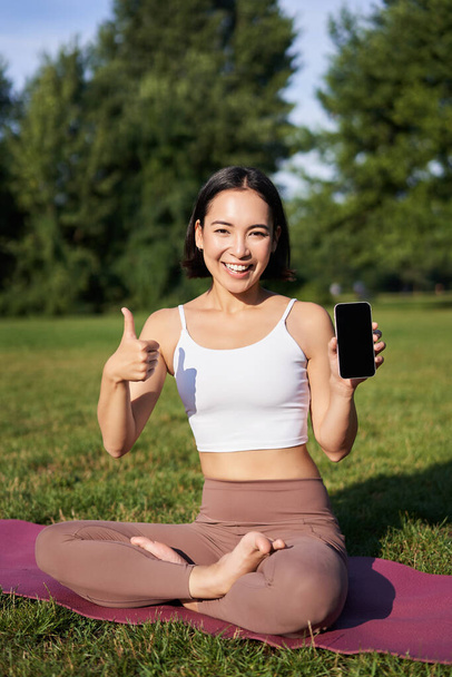 Vertical shot of asian woman shows thumbs up, recommending yoga training online, meditation app, doing exercises on fresh air in park, showing smartphone screen. - Photo, Image