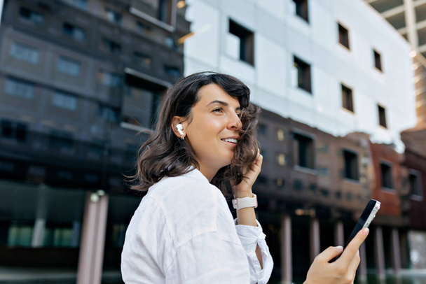De cerca retrato de perfil de romántica mujer de pelo oscuro bastante en los auriculares está sosteniendo el teléfono inteligente y sonriendo sinceramente  - Foto, Imagen