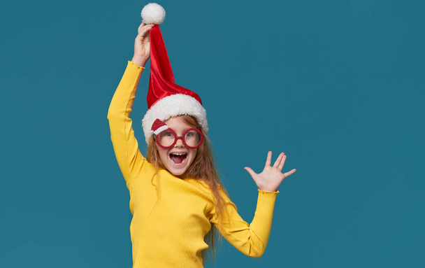 Funny little girl in a Santa Claus hat and party glasses on a turquoise background. The child smiles in surprise with his mouth wide open. wow funny face - Fotó, kép