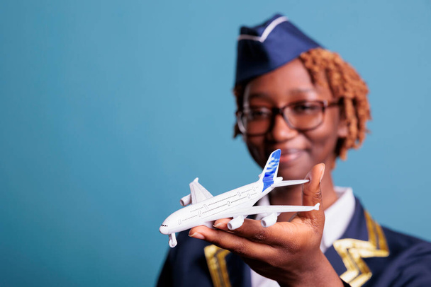 Aircraft attendant in crew uniform looking at model airplane, holding it in his hands with a smile. Female professional flight assistant with commercial toy airplane in studio shot. - Photo, Image