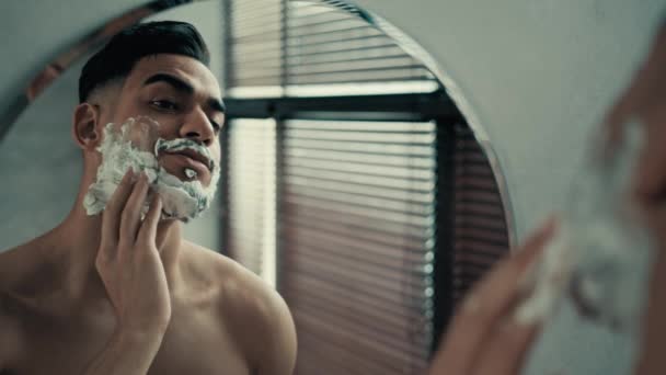Hispanique Latina Indien barbu mâle non rasé avec soies millénaires homme barbouiller mousse blanche gel de rasage cosmétique frottis sur le visage joues barbe regarder miroir réflexion se préparer à raser les poils du visage - Séquence, vidéo