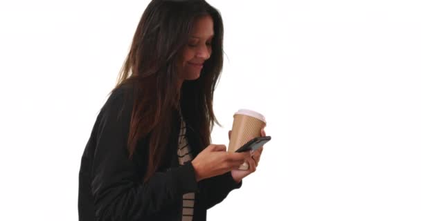 Hymyilevä nainen pommikonetakissa pitelemässä kahvia tekstaillessaan kännykkä valkoisella taustalla. Millennial nainen messaging ystävä älypuhelin studiossa copyspace. 4 k. - Materiaali, video