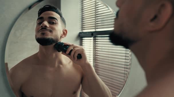 Lähikuva mies heijastus peilistä kylpyhuoneessa parranajo harjakset kasvoilla parranajo kotona. Intialainen latino komea 30s kaveri parrakas latino mies käyttää sähköinen parranajokone sähköinen trimmeri parranajo - Materiaali, video