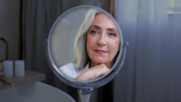 Mosolygó gyönyörű női arc 50-es évekbeli középkorú érett nő élvezze arc bőr nézi tükör tükröződés kényeztető. Öreg, száraz bőr szépségápolási eljárások kozmetikai anti age plasztikai sebészet - Felvétel, videó
