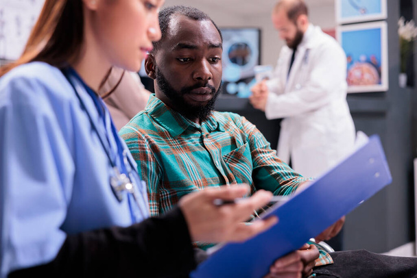 Közelkép az afro-amerikai férfiról, aki átnézi az orvosi feljegyzéseket az ázsiai női kórházi alkalmazottakkal együtt. Az orvos kezelést javasol fiatal férfi betegnek a szanatórium előcsarnokában. - Fotó, kép