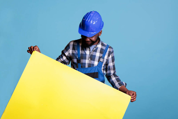 Οικοδόμος κοιτάζοντας κάτω μεγάλο κίτρινο πίνακα που κρατά ψηλά για τη διαφήμιση με την έννοια της κατασκευαστικής βιομηχανίας. Αφροαμερικάνος με στολή και σκληρό καπέλο παρουσιάζοντας πίνακα με διαφημιστικό χώρο αντιγραφής. - Φωτογραφία, εικόνα