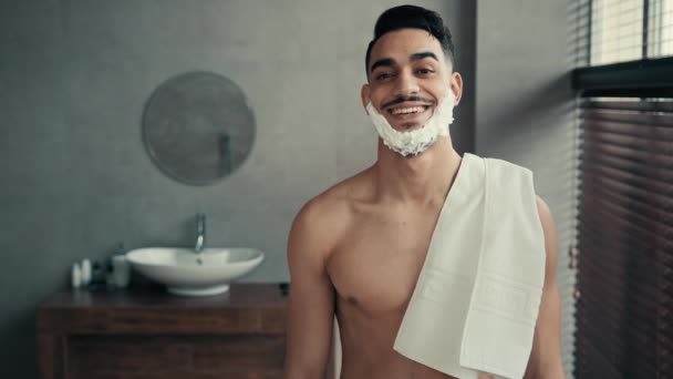 Vicces férfi portré a fürdőszobában Latina fickó mosoly pózol előtt borotvált szakáll. Spanyol indián mosolygós boldog szexi férfi szakállas fickó fehér szappan hab szakáll borotválkozás gél gazdaság kád törölköző higiénia - Felvétel, videó
