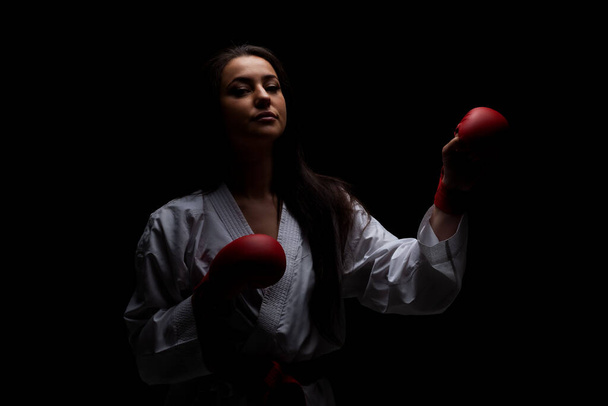 girl exercising karate punch wearing kimono and red gloves against black background - Foto, imagen