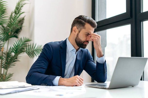 Frustrated overworked caucasian businessman, entrepreneur, product manager, programmer, sits at a desk in the office, massaging the bridge of his nose with his eyes closed, exhausted from long work - 写真・画像
