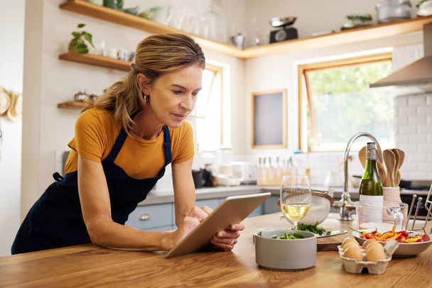 Woman, tablet and kitchen for online recipe, browsing or research in cooking or baking at home. Female with apron checking instructions on the internet for meal or ingredients on touchscreen indoors. - Foto, Bild